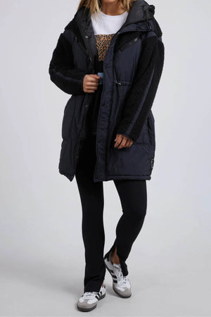A person in a dark Foxwood Longline Anorak with faux fur sleeves, white shirt, black leggings, and black and white sneakers stands against a neutral backdrop. Made for outdoor adventure from durable nylon polyester blend, they have their hands in the coat pockets, face unseen.