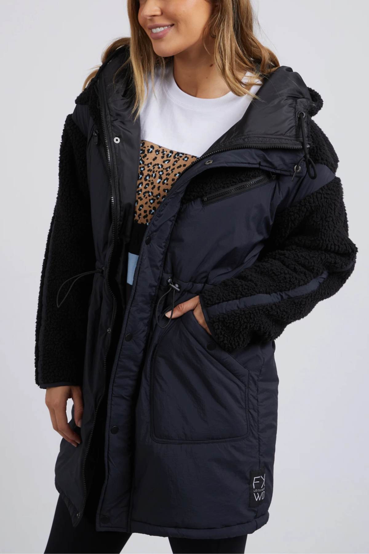 A person wearing the Foxwood Longline Anorak, a black hooded parka with teddy fabric on sleeves and shoulders, ideal for outdoor adventures. Their hand is in one pocket while a white shirt with a leopard print band peeks out. The nylon polyester blend ensures durable style and function in any weather. .