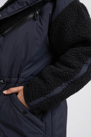 Close-up of someone in the Foxwood Longline Anorak, a dark winter coat made for outdoor adventures, featuring a nylon-polyester blend with wool-like textured sleeves and sides. It has a front zipper pocket, snap button detail, with one hand in the pocket.