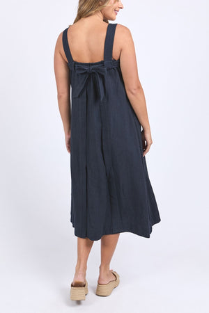 A woman with light brown hair faces away, wearing a sleeveless Foxwood Lourdes Dress in dark blue with wide straps, an open back featuring a large bow, and wedge sandals, set against a plain white background.