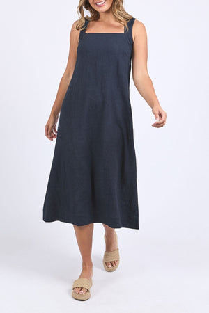 A woman models the Foxwood Lourdes Dress, a sleeveless, navy blue, ankle-length linen dress with a square neckline, paired with beige wedge sandals, against a plain white background.