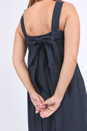 Someone stands with their hands clasped behind them, wearing the Foxwood Lourdes Dress—a navy sleeveless linen dress with wide straps and a tied bow at the back.