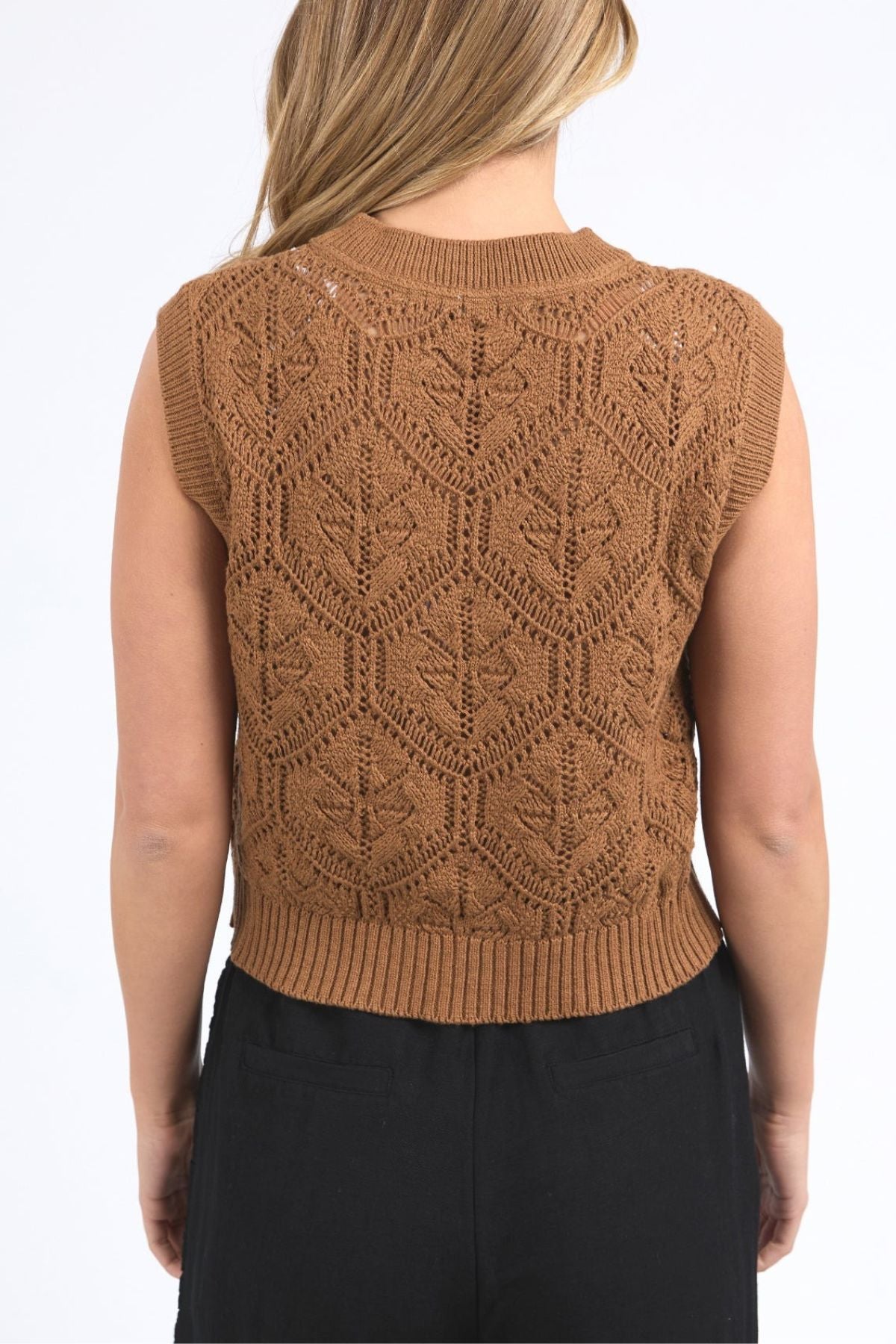 A person shown from the back wears the sleeveless Foxwood Luna Knit Vest in brown, featuring a leaf-like pointelle pattern and ribbed edges, paired with high-waisted black pants.