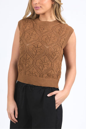 Woman wears the Foxwood Luna Knit Vest—a sleeveless, brown pointelle knit top with a geometric pattern—paired with high-waisted black pants. The photo focuses on her upper body; her head is partially out of frame.