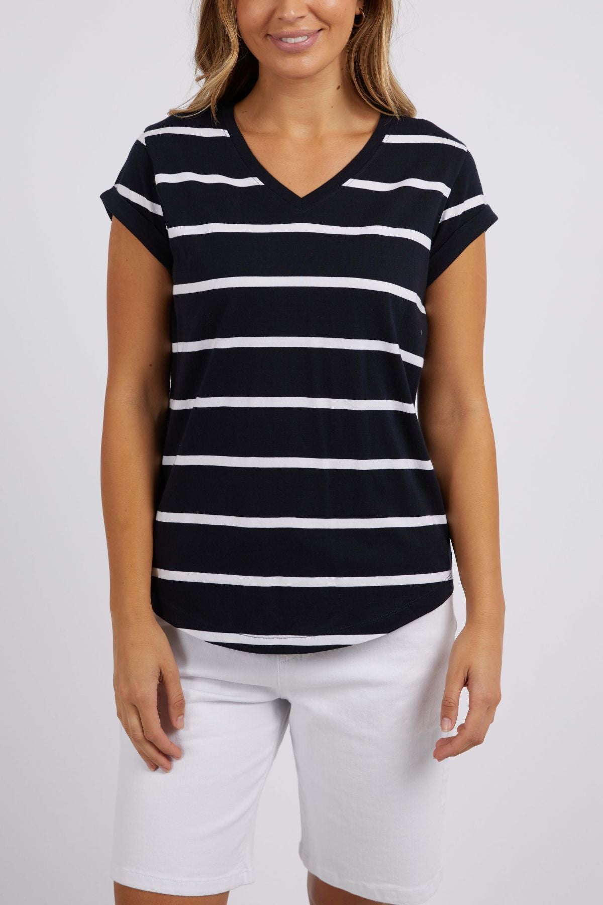 A person wearing the Foxwood Manly Stripe Vee Tee, a V-neck T-shirt made of soft Pima cotton jersey with black and white yarn-dyed stripes, paired with white shorts stands against a plain background. The T-shirt features short sleeves and a casual fit.