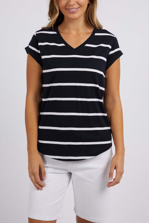 A person wearing the Foxwood Manly Stripe Vee Tee, a V-neck T-shirt made of soft Pima cotton jersey with black and white yarn-dyed stripes, paired with white shorts stands against a plain background. The T-shirt features short sleeves and a casual fit.