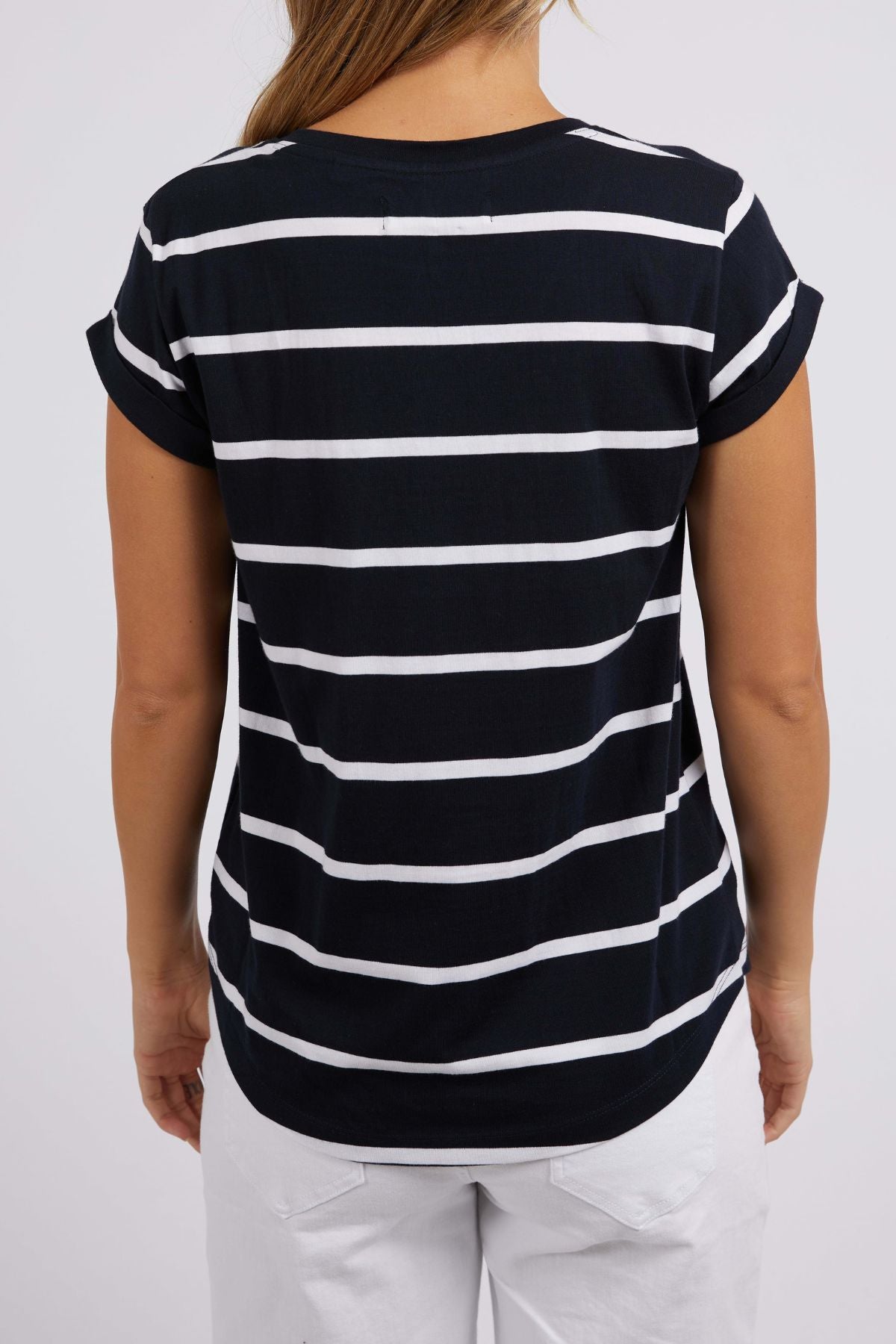 The individual is donning the Foxwood Manly Stripe Vee Tee, a black and white yarn-dyed striped T-shirt crafted from soft Pima cotton jersey, featuring short sleeves. Positioned with their back to the camera, they complete the look with crisp white pants set against a plain, light-colored backdrop.