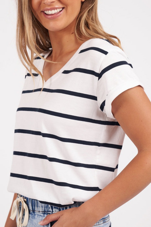 A woman in a Foxwood Manly Stripe Vee Tee smiles, posing with one arm on her waist. The vee neckline complements her long blonde hair, and the outfit includes casual blue jeans. The background is plain white.