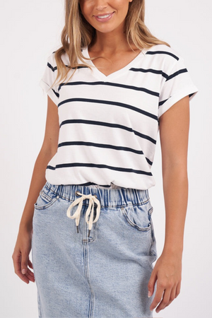 A person wearing the Foxwood Manly Stripe Vee Tee, a white and black yarn-dyed stripe T-shirt made of soft Pima cotton jersey, and a light denim skirt with a drawstring stands against a plain background.