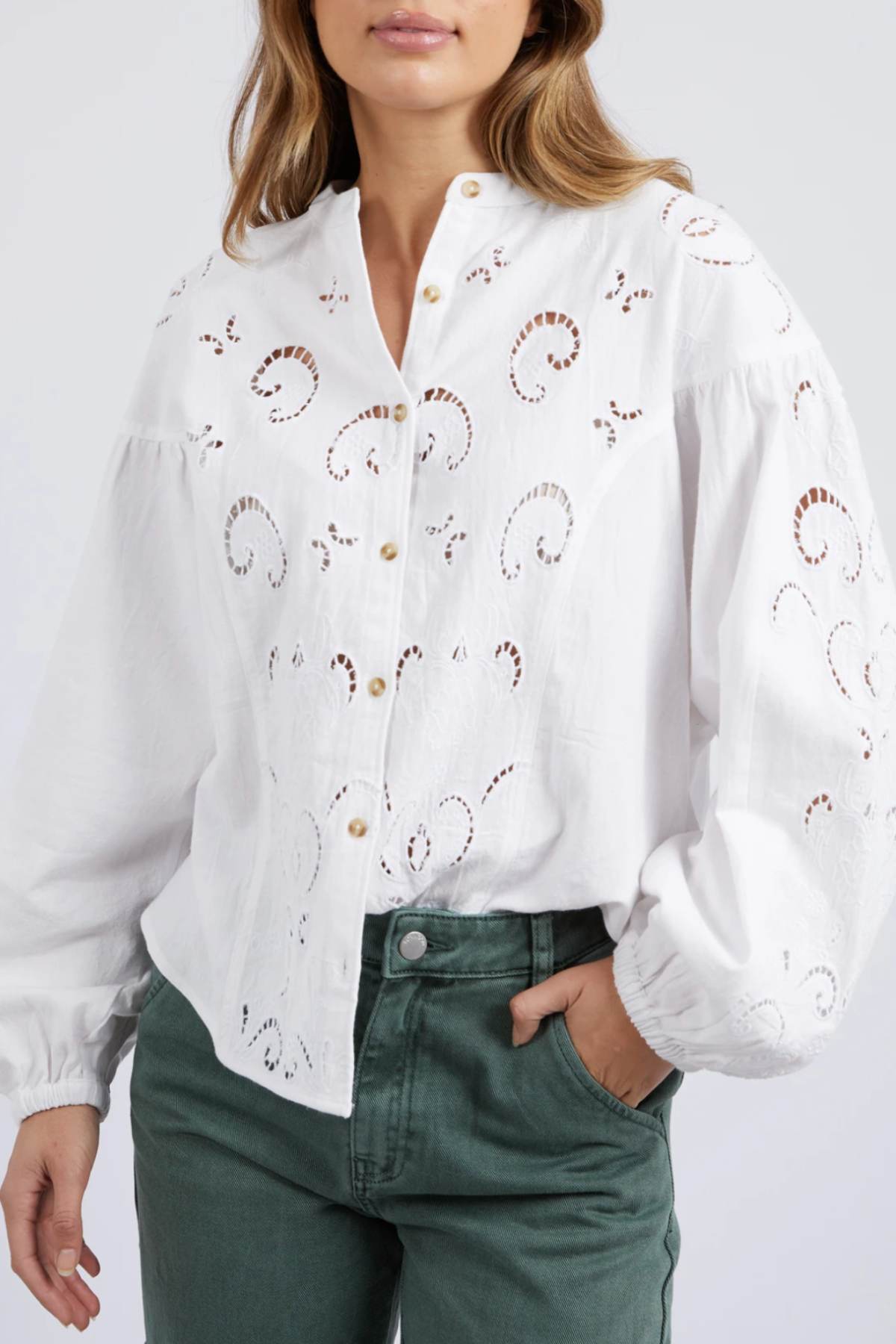 A woman wears the Foxwood Marlow Blouse featuring eyelet-inspired embroidery and gold buttons, paired with high-waisted green pants; her hand rests in her pocket.