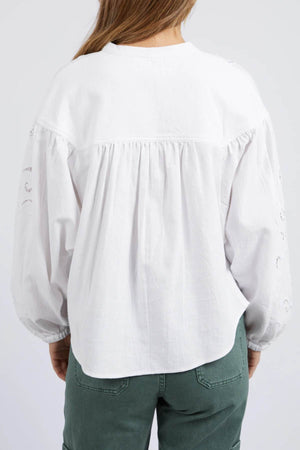 A person with long brown hair is seen from behind, wearing a loose-fitting Foxwood Marlow Blouse with gathered detailing and eyelet-inspired embroidery on the sleeves, paired with green pants.