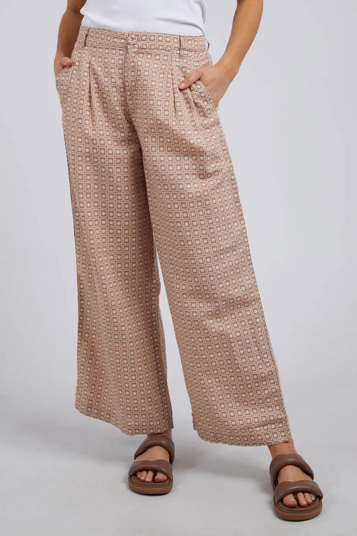 The person wears Foxwood Maya Geo Pant, showcasing a chic geometric pattern on high-waisted beige wide-leg pants. Made from a linen/cotton blend, their hands in pockets complete the look with a white top and brown sandals against a plain gray backdrop.