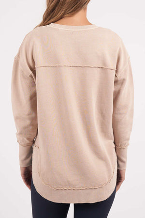 A person with long hair, seen from the back, is wearing an oversized beige Foxwood Medalion Crew sweater that features visible seams and raw edge detailing. The sweater offers a relaxed, casual fit and is paired with dark-colored pants. The background is plain white.
