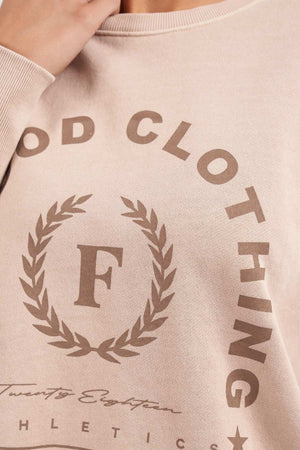 Close-up of a person wearing the Foxwood Medalion Crew, a beige sweatshirt made from 100% cotton. It features the text "Food Clothing" in a circular design and the letter "F" inside a laurel wreath. Below the laurel wreath, there are additional texts including "Twnty Eight Athletics." The sweatshirt also has raw edge detail for added style.