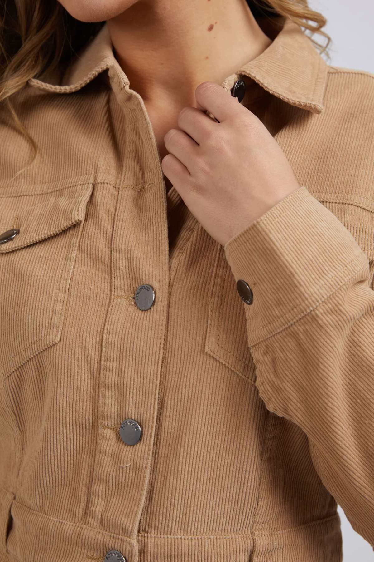 A person wearing the tan Foxwood Montana Cord Dress made of 100% cotton with metallic buttons holds the collar, with the image focusing on the upper torso to showcase the garment’s rich corduroy texture and detailed fabric.