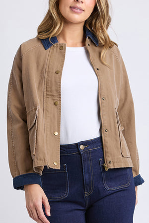 A woman wears the Foxwood Colwood Jacket, a sporty tan jacket with brass buttons, navy corduroy collar and cuffs, large front pockets, and a slightly cropped fit over a white top and high-waisted dark blue jeans.