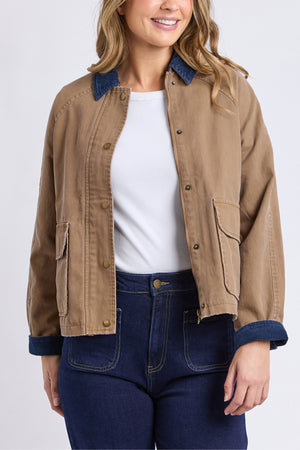 A woman wears the Foxwood Colwood Jacket in tan with a sporty look, blue contrast collar and cuffs, styled over a white shirt and high-waisted dark blue jeans. The image shows her from shoulders to mid-thigh.