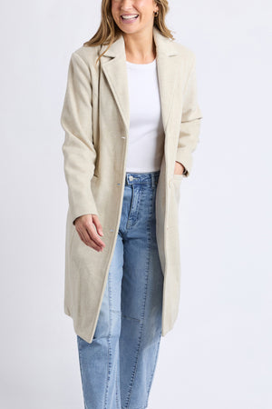 A woman wears the Foxwood Nelson Coat in light beige over a white top and light blue high-waisted jeans, smiling with one hand in her coat pocket and the other slightly raised, standing against a plain background.