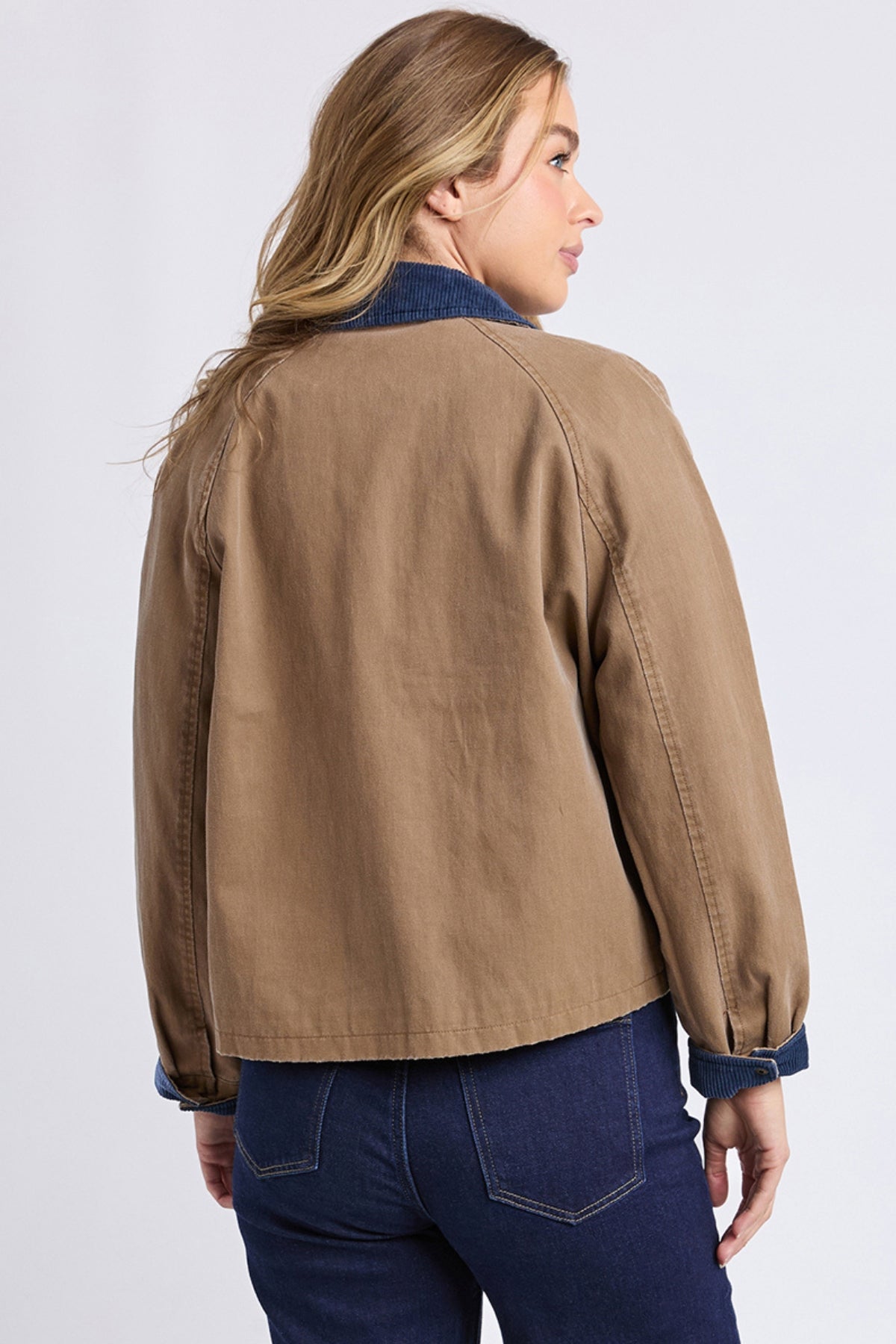 A person with long, light brown hair is wearing a tan Foxwood Colwood Jacket featuring a blue contrast collar and cuffs, paired with dark blue jeans, facing away and slightly turned to the side against a plain light background.