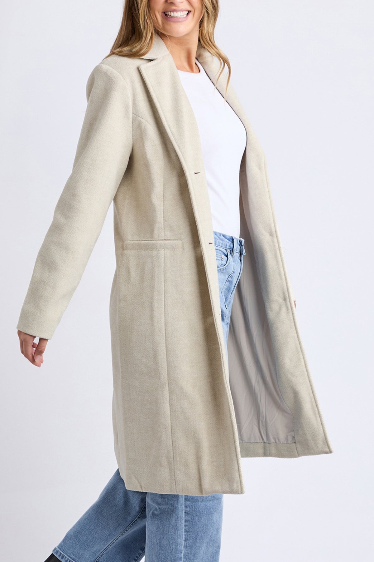 A woman smiles in a light beige Foxwood Nelson Coat, layered over a white top and blue jeans, standing against a plain light background.