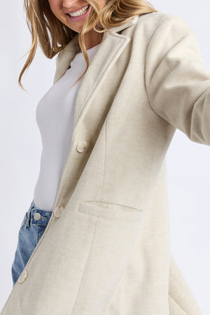 A woman with light brown hair smiles and takes a selfie, wearing the cream-colored Foxwood Nelson Coat over a white top and blue jeans.