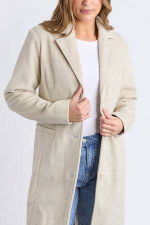 A person wears a light beige Foxwood Nelson Coat over a white shirt and blue jeans, standing against a plain background with hands holding the front of the mid-length coat.