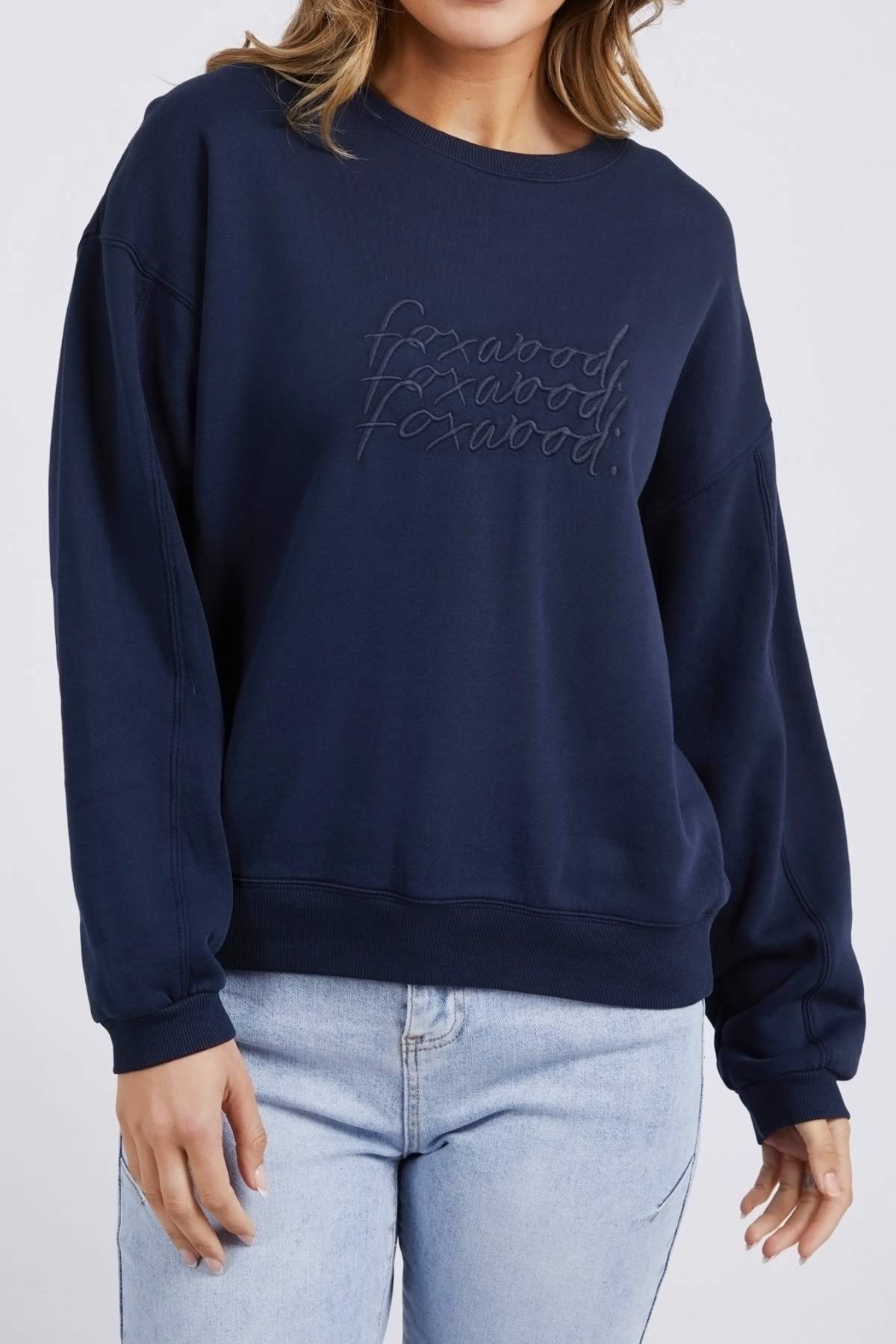 A person is wearing the Foxwood Nimble Crew in navy blue, showcasing playful front chest embroidery. The 100% cotton sweatshirt features long, relaxed-fit sleeves and is paired with light blue jeans. The person's face is not visible in the image.