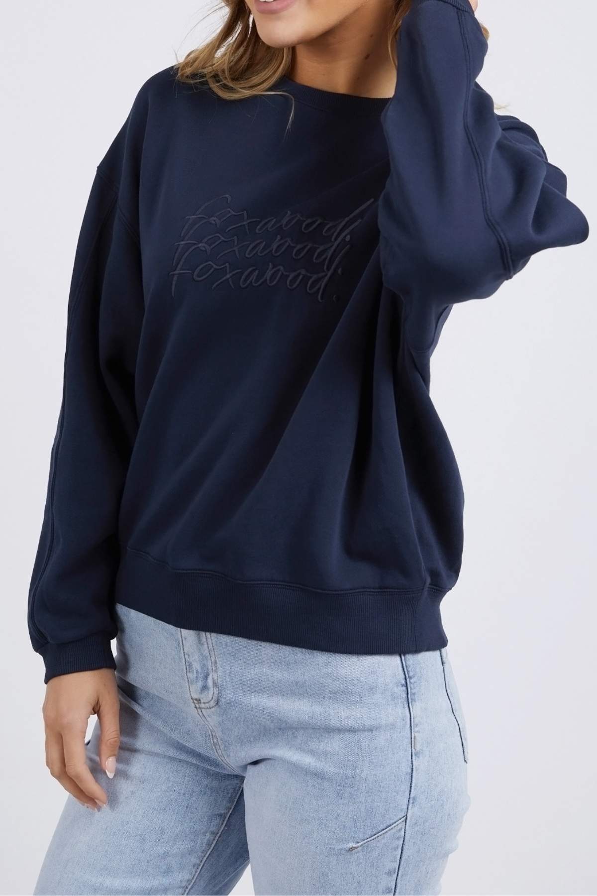 A person wearing the Foxwood Nimble Crew, a navy blue 100% cotton sweatshirt with playful front chest embroidery, and light blue jeans is posing with one hand touching their hair and the other hand resting by their side. The relaxed fit top stands out against the plain white background.