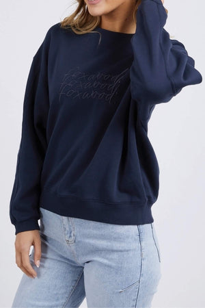 A person wearing the Foxwood Nimble Crew, a navy blue 100% cotton sweatshirt with playful front chest embroidery, and light blue jeans is posing with one hand touching their hair and the other hand resting by their side. The relaxed fit top stands out against the plain white background.