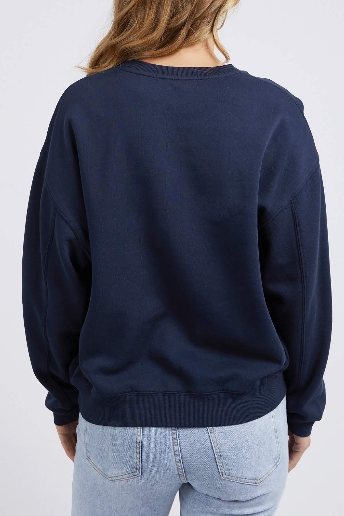 A person with long brown hair stands facing away from the camera, wearing a dark blue Foxwood Nimble Crew top and light blue jeans. The background is a plain white wall.