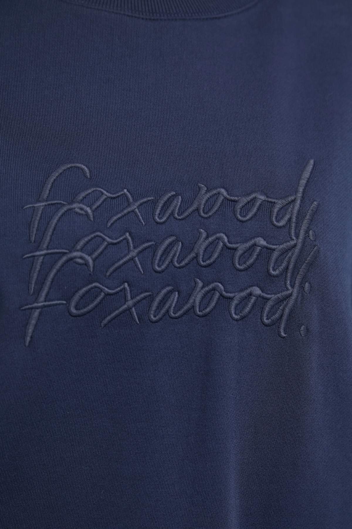 A close-up of a navy blue fabric from the Foxwood Nimble Crew relaxed fit top shows the word "foxwood" stitched in a repetitive, overlapping pattern in the same navy blue color, creating a subtle textured effect. The playful front chest embroidery adds charm to this 100% cotton piece.