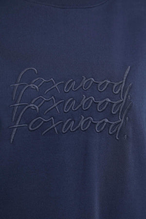 A close-up of a navy blue fabric from the Foxwood Nimble Crew relaxed fit top shows the word "foxwood" stitched in a repetitive, overlapping pattern in the same navy blue color, creating a subtle textured effect. The playful front chest embroidery adds charm to this 100% cotton piece.