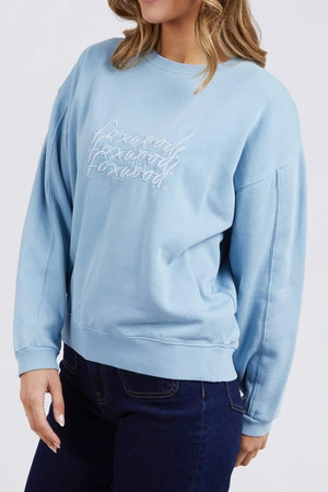 A woman is wearing a Foxwood Nimble Crew sweatshirt in light blue, featuring long sleeves and white embroidered text on the front that reads "Forward Forward Forward." This relaxed fit top, made from 100% cotton, is paired with dark blue jeans. The background is plain white.