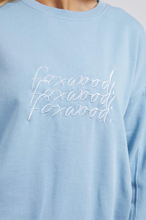 Close-up of a person wearing a light blue Foxwood Nimble Crew sweatshirt with white embroidered text. Made from 100% cotton for ultimate comfort, this relaxed-fit top ensures ease of movement. The text seems to repeat and overlap, making it difficult to discern the word.