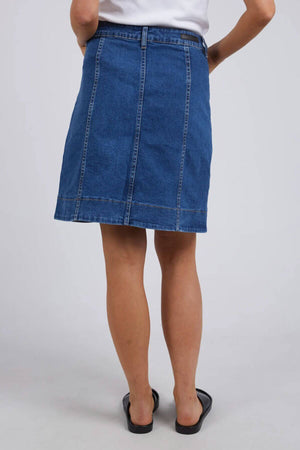 Someone stands facing away from the camera against a plain light background, wearing a blue Foxwood Nolan Skirt with contrast stitching, paired with a white top and black sandals.