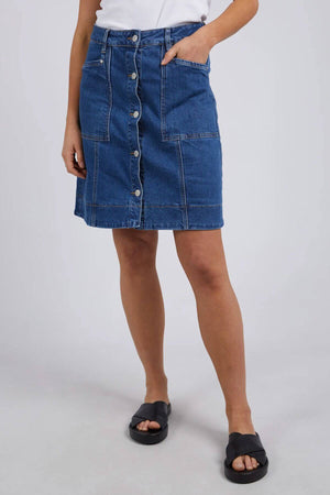 A person wearing a white top, the Foxwood Nolan Skirt—a blue denim A-line skirt—and black slide sandals stands with one hand in a pocket against a plain light background.