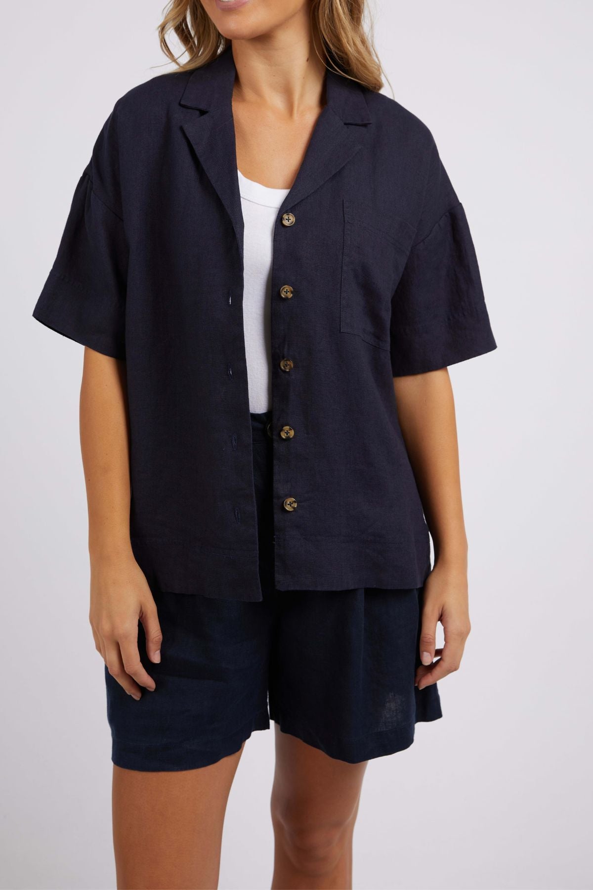 A person is dressed in a Foxwood Oakleigh Shirt in navy blue, recognized for its moisture-wicking features, layered over a white top and complemented by navy blue shorts. The backdrop is a plain light gray.