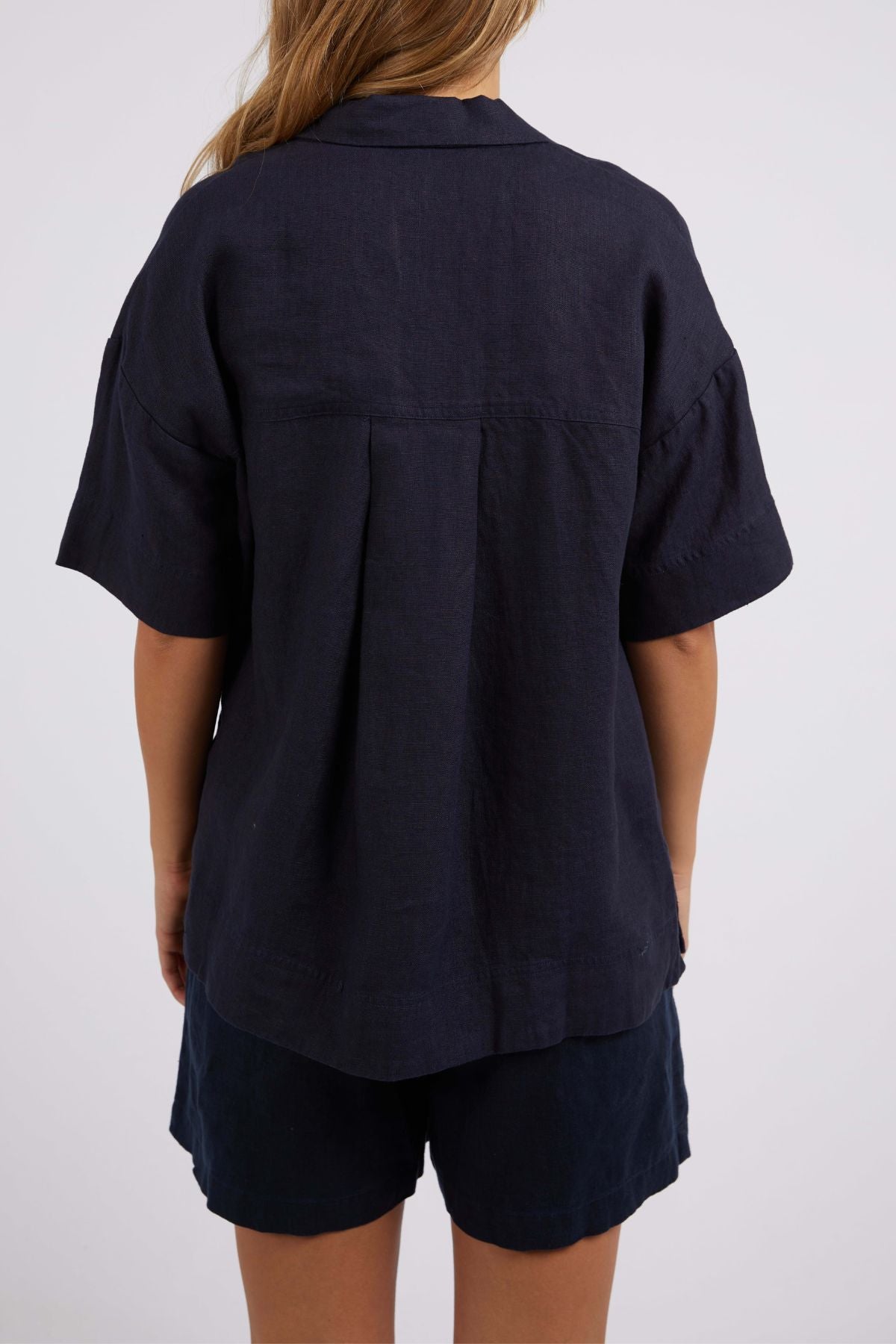 A person with long hair is dressed in a dark blue, short-sleeve Foxwood Oakleigh Shirt paired with matching shorts, highlighting the shirt's moisture-wicking features, as they face away from the camera set against a simple backdrop.