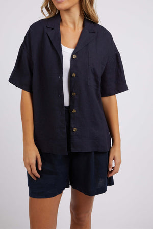 A person is dressed in a Foxwood Oakleigh Shirt in navy blue, recognized for its moisture-wicking features, layered over a white top and complemented by navy blue shorts. The backdrop is a plain light gray.