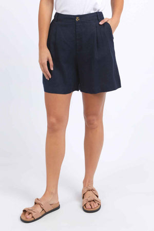 A person wearing Foxwood Oakleigh Shorts, paired with a white shirt, stands against a plain background. They complete the look with beige sandals featuring bows.