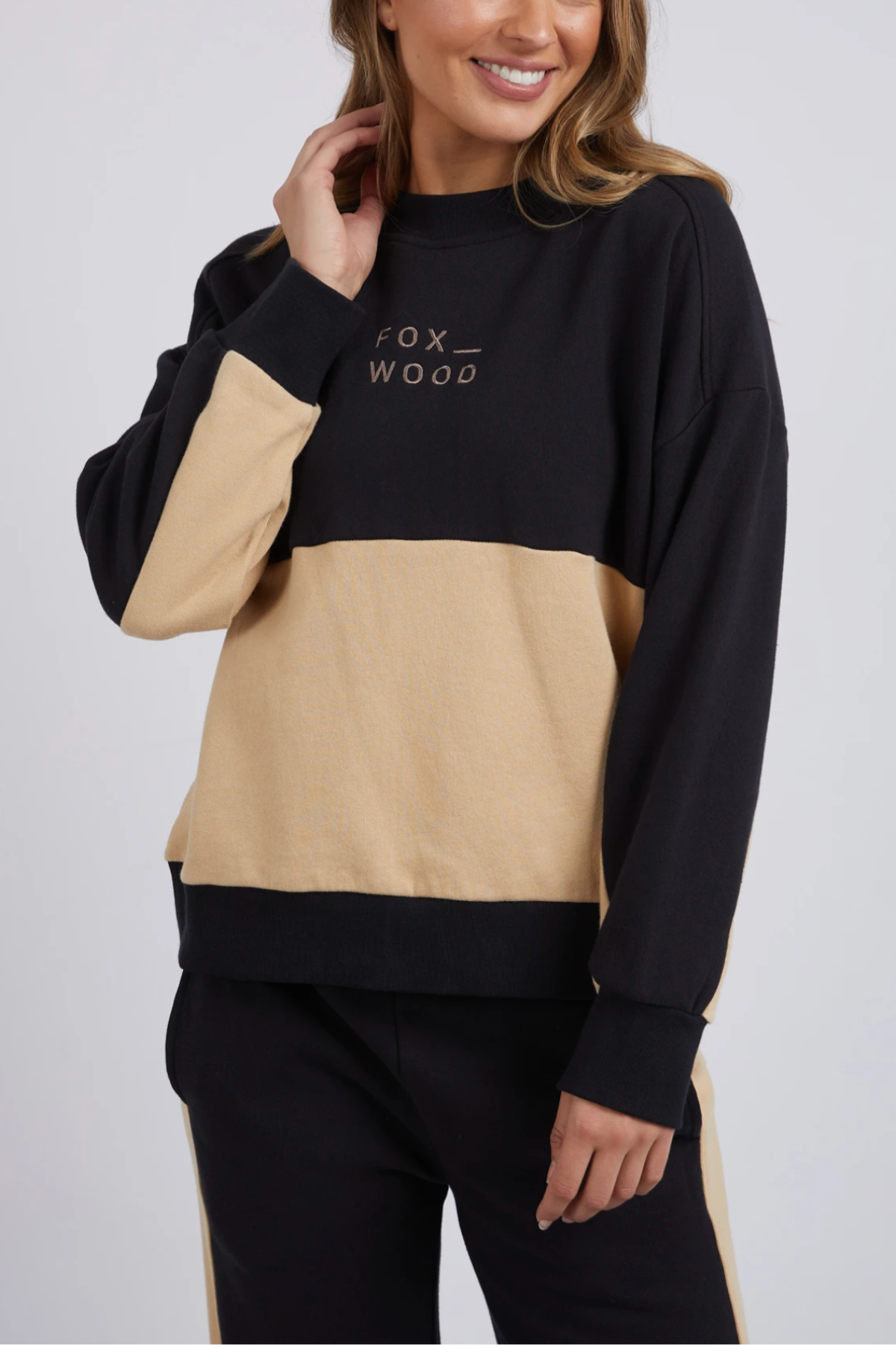 A person wearing a black and beige Foxwood Offhand Crew sweatshirt with "FOX WOOD" printed on it is smiling, one hand in their long hair. Made from cotton fleece, this outfit offers lounge comfort and features coordinating color-block pants.