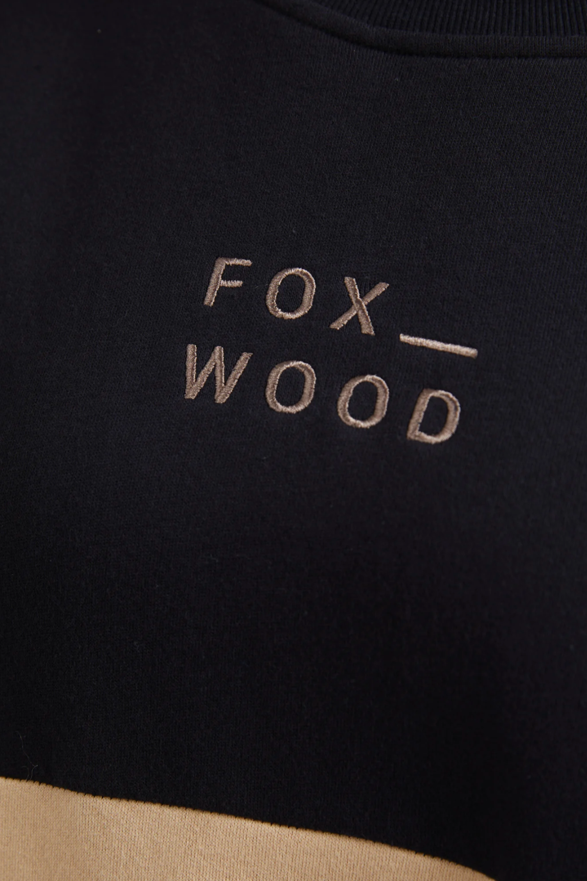 Close-up of a black cotton fleece fabric embroidered with "FOX WOOD" in beige thread. Its smooth texture boosts comfort, with centered embroidery reflecting the Foxwood Offhand Crew style essence.