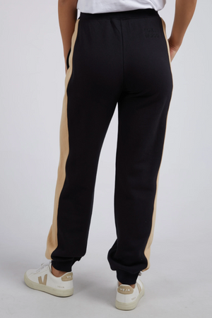Person wearing black Foxwood Offhand Pant with beige side stripes, a white 100% cotton fleece T-shirt, and white Veja sneakers stands against a light background. The back view highlights the lounge comfort of the Offhand Pant.