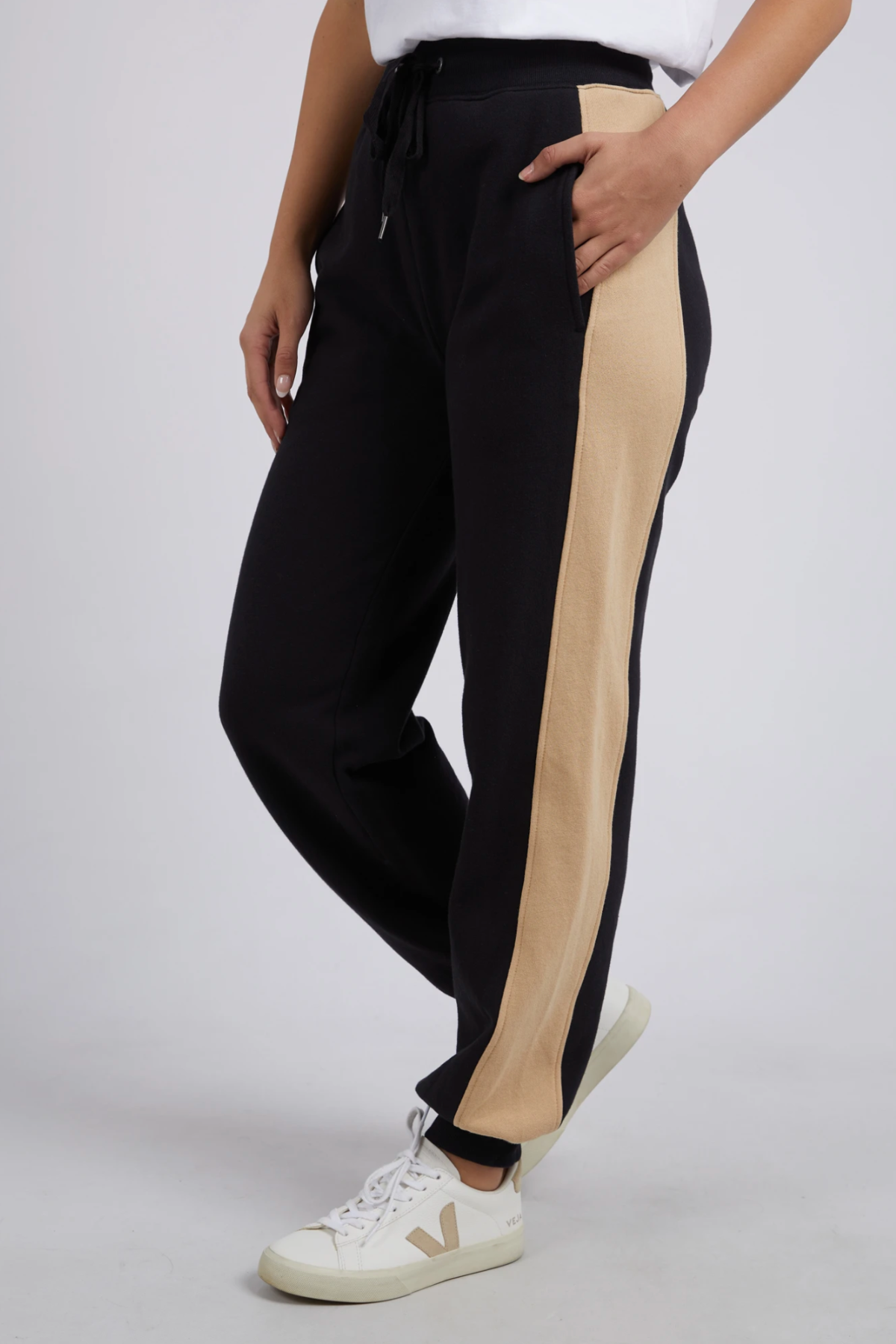 Someone wearing Foxwood Offhand Pant joggers with wide beige side stripes, white sneakers, and a white top stands with one hand in their pocket, highlighting the pants’ 100% cotton fleece comfort against a plain background.