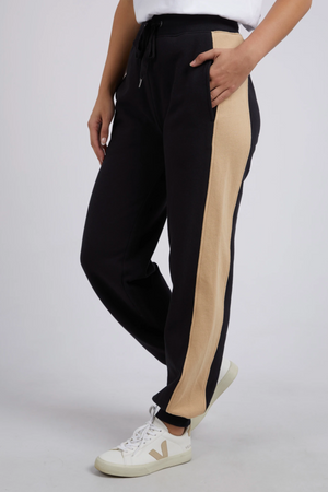 Someone wearing Foxwood Offhand Pant joggers with wide beige side stripes, white sneakers, and a white top stands with one hand in their pocket, highlighting the pants’ 100% cotton fleece comfort against a plain background.