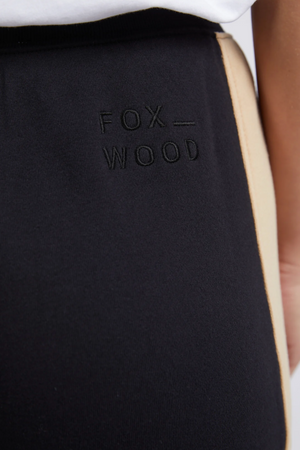 The Foxwood Offhand Pant features a beige side stripe, "FOX WOOD" embroidery on the hip, and a relaxed fit cotton fleece design. Shown paired with a white shirt.