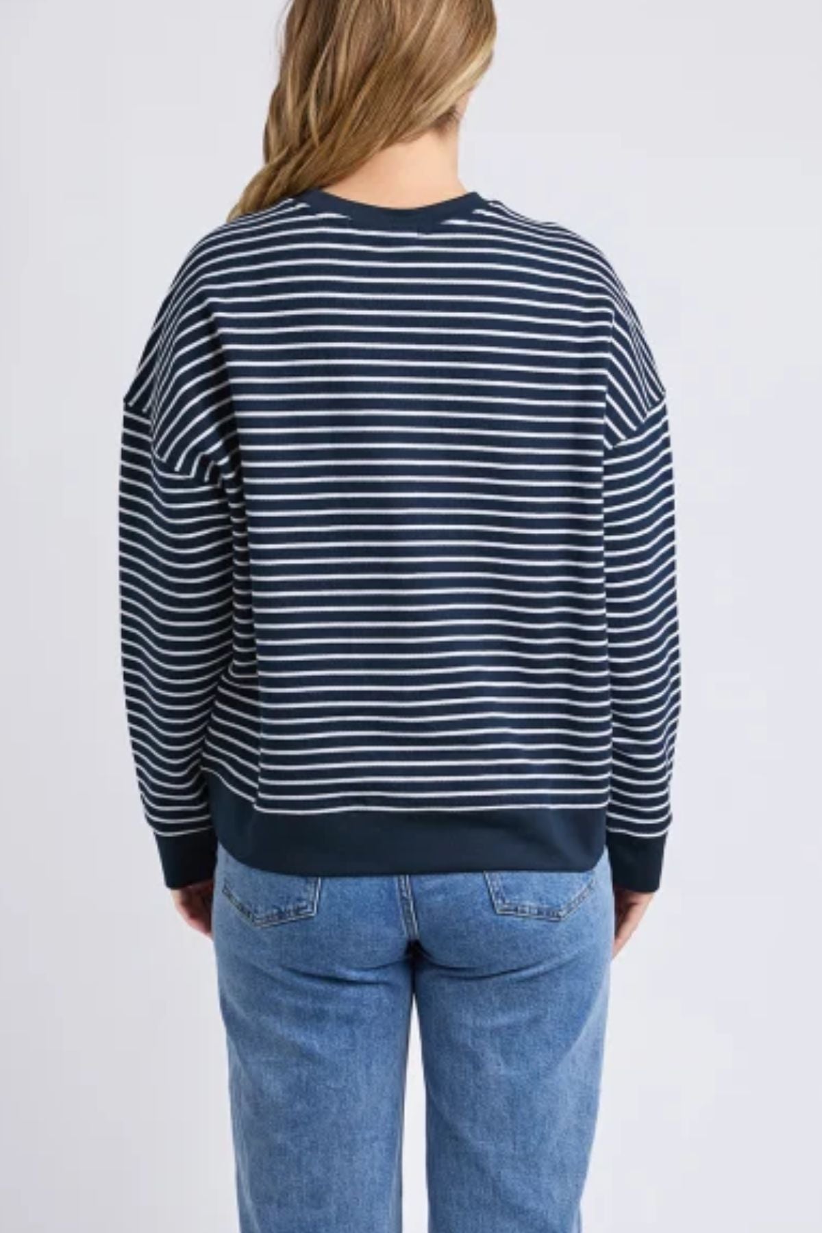 A person with long, light brown hair is seen from behind in the Foxwood Original Stripe Crew jumper and blue jeans, showcasing effortless winter style against a plain light background.