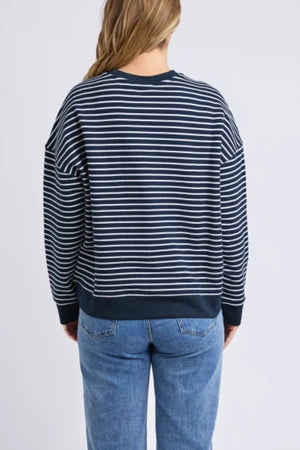 A person with long, light brown hair is seen from behind in the Foxwood Original Stripe Crew jumper and blue jeans, showcasing effortless winter style against a plain light background.