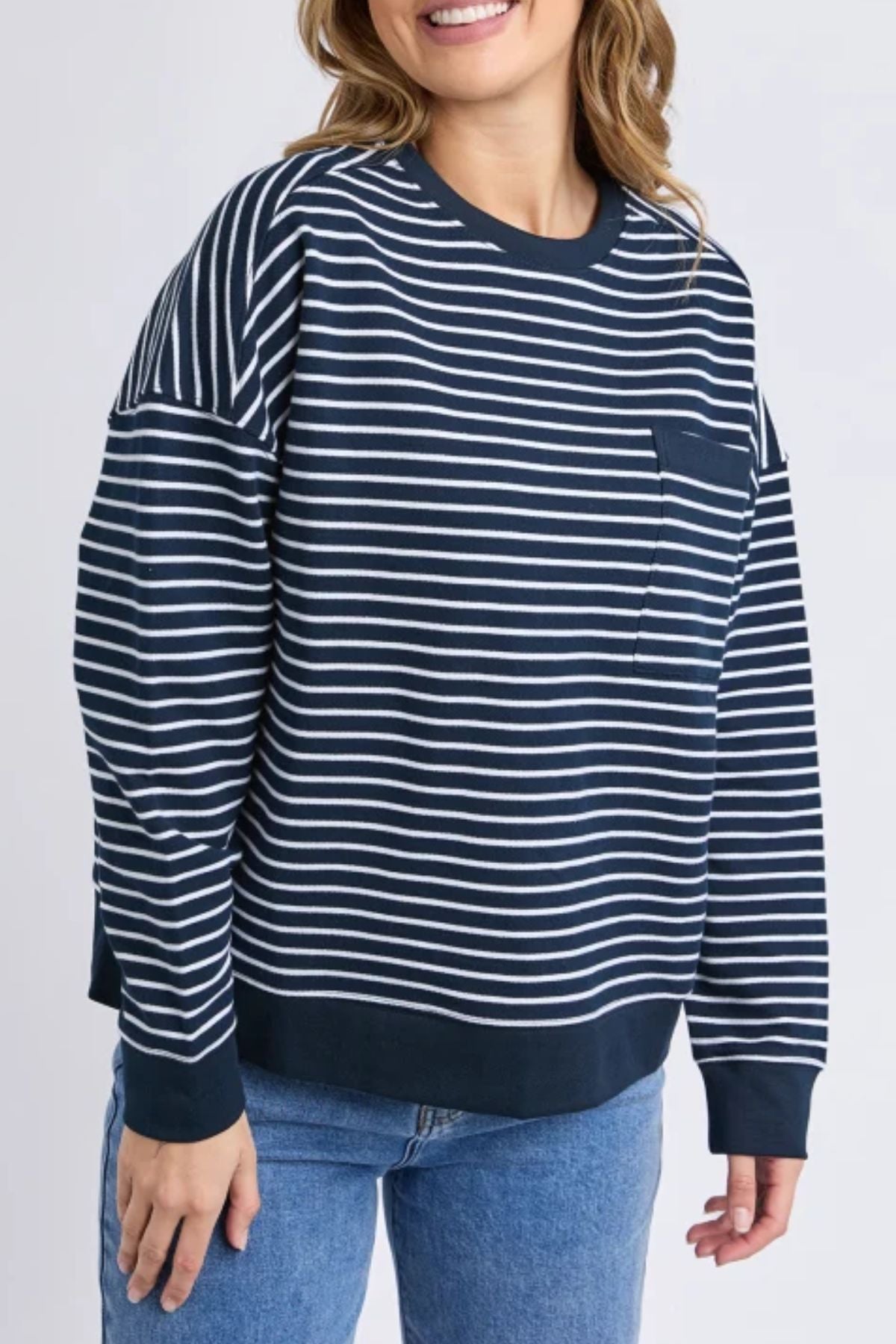 A woman wears the Foxwood Original Stripe Crew, a navy and white relaxed fit jumper with a chest pocket, paired with blue jeans. She smiles against a neutral background—perfect for effortless winter style.