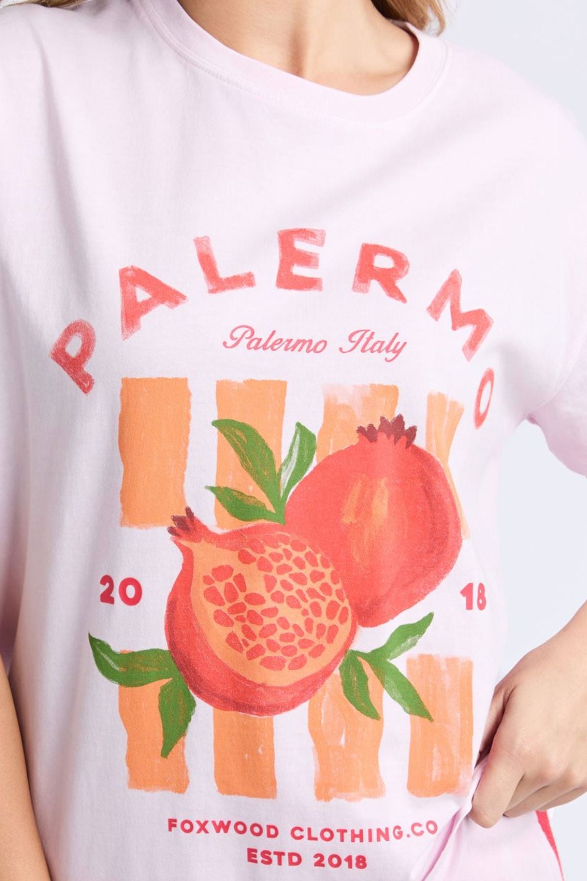 Someone wears the Foxwood Palermo Tee in relaxed-fit light pink, featuring two pomegranates, orange stripes, and text: "PALERMO," "Palermo Italy," "2018," and "FOXWOOD CLOTHING CO ESTD 2018" in a vibrant Foxwood print.
